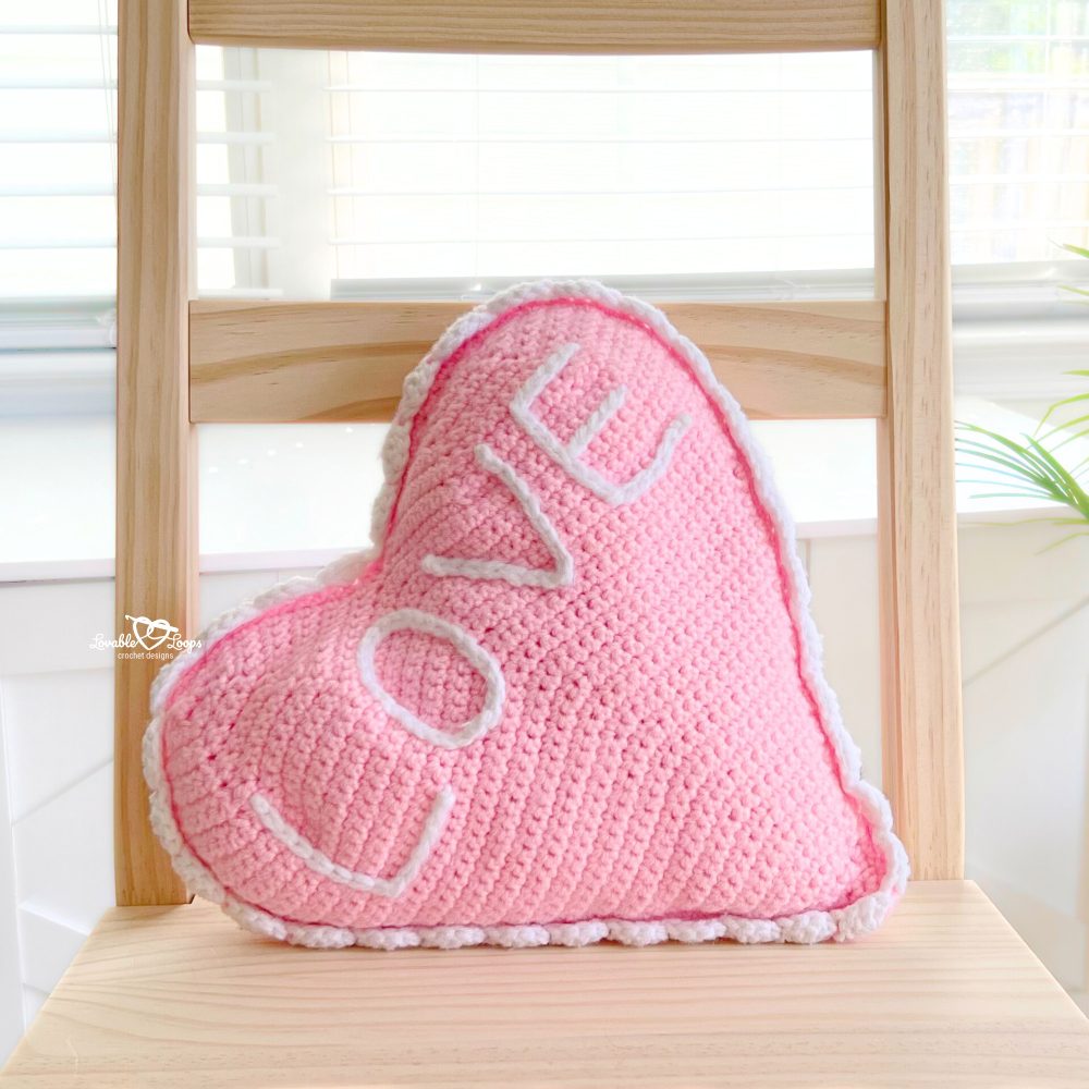 Close-up of pink crochet heart pillow with scalloped white border and “LOVE” stitched on front, placed on a light wood chair near a window. Handmade gift or decor idea.