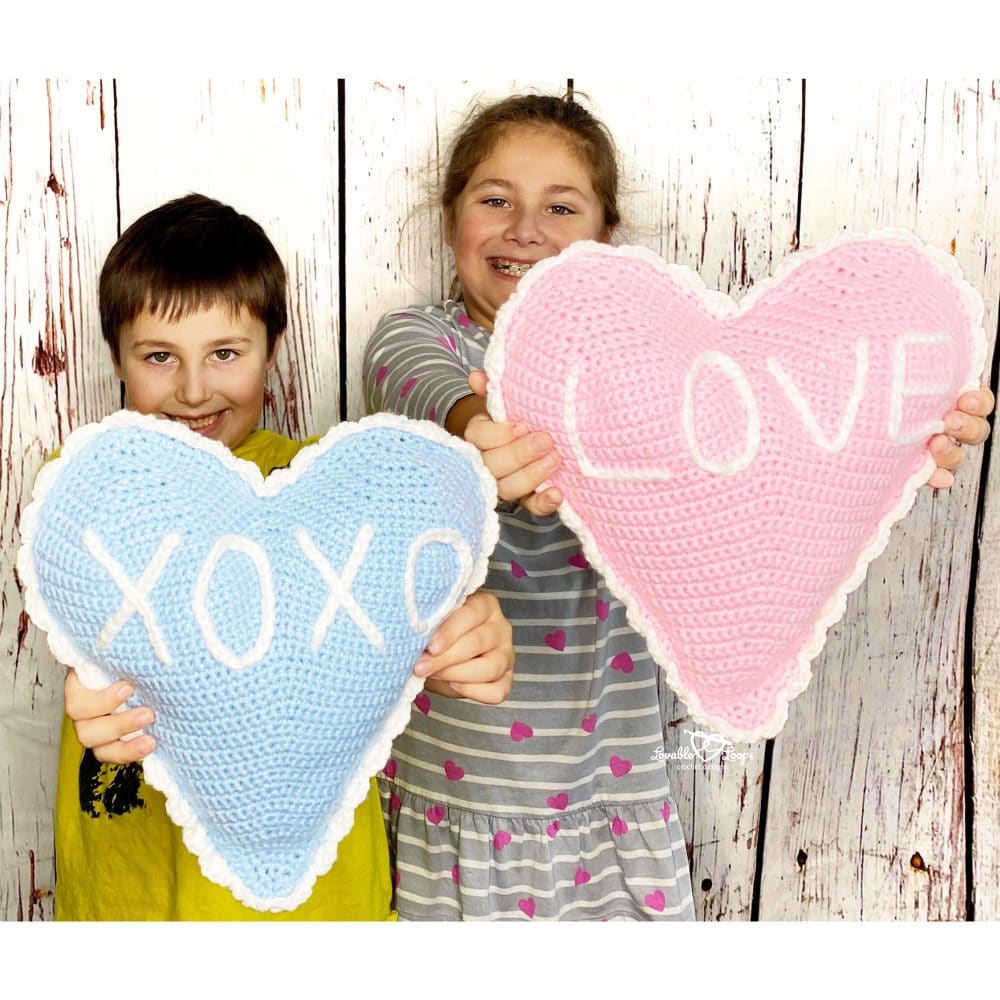 Smiling kids holding heart-shaped crochet pillows—one blue with “XOXO,” one pink with “LOVE.” White ruffled border around each pillow. Fun DIY Valentine’s Day photo.