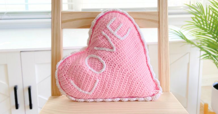 Pink crochet heart pillow with white border and surface slip-stitched “LOVE” in white yarn, displayed on a wooden chair in front of a bright window. Cozy handmade Valentine’s Day home decor.