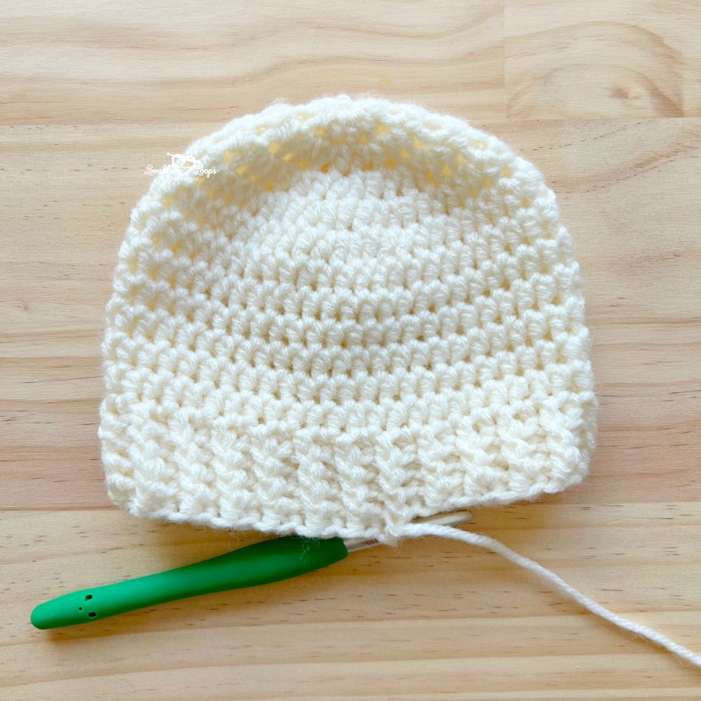 Cream crochet baby hat in progress, with ribbed brim and a green crochet hook inserted into the working yarn, placed on a light wooden surface.
