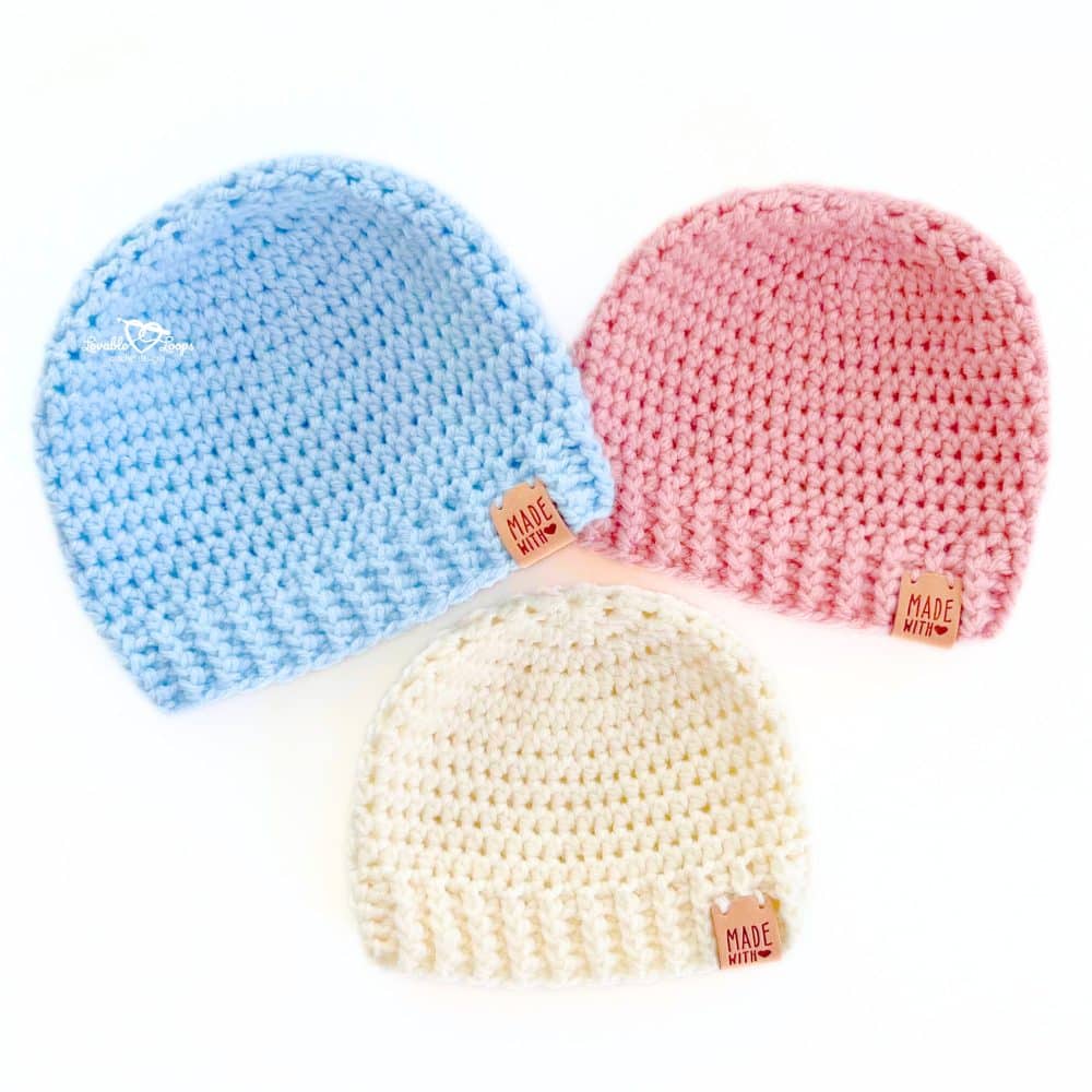 Three handmade crochet baby hats in blue, pink, and cream yarn displayed in a flat lay. These beginner-friendly baby beanies use basic stitches and are part of a free crochet baby hat pattern.