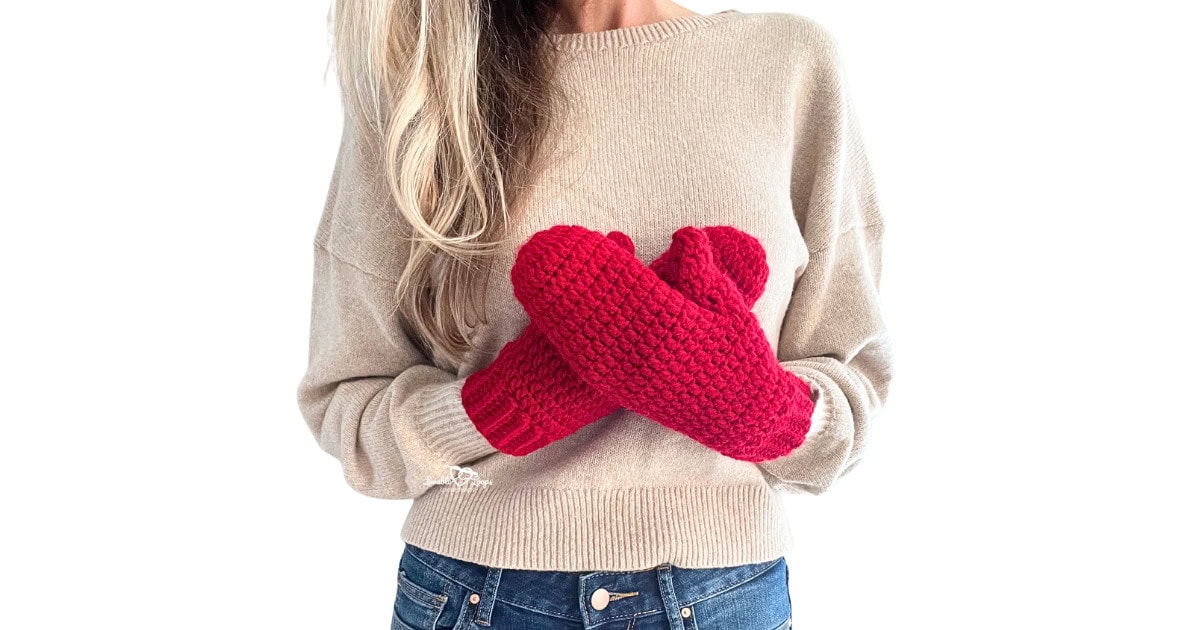 Woman wearing bright red crochet mittens with ribbed cuffs, crossing hands in front of a beige sweater. These handmade winter gloves are part of a free easy crochet mittens pattern that’s beginner-friendly and cozy for cold weather.