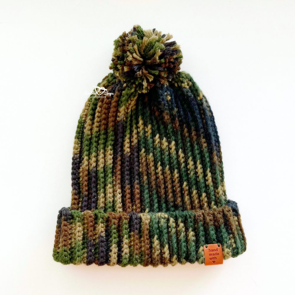 Camo-style crochet men’s beanie displayed on a white surface with a folded brim and a yarn pom pom on top. An easy crochet hat pattern for men with a ribbed texture, worked flat in rows.
