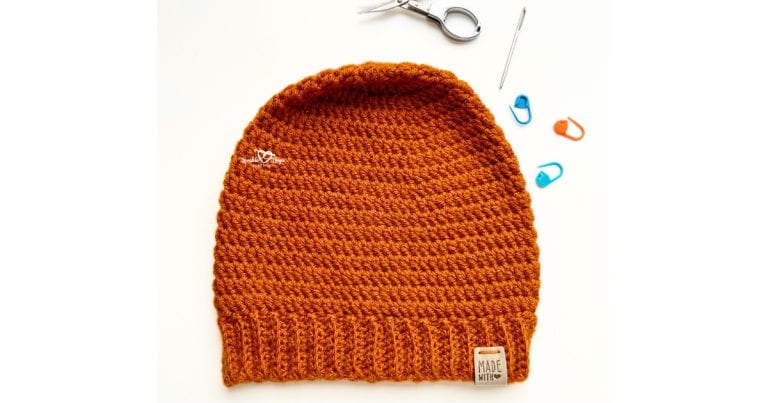 Flat lay of a burnt orange men’s crochet hat with ribbed brim, surrounded by scissors, a yarn needle, and stitch markers. This free & easy beanie crochet pattern is perfect for beginners.