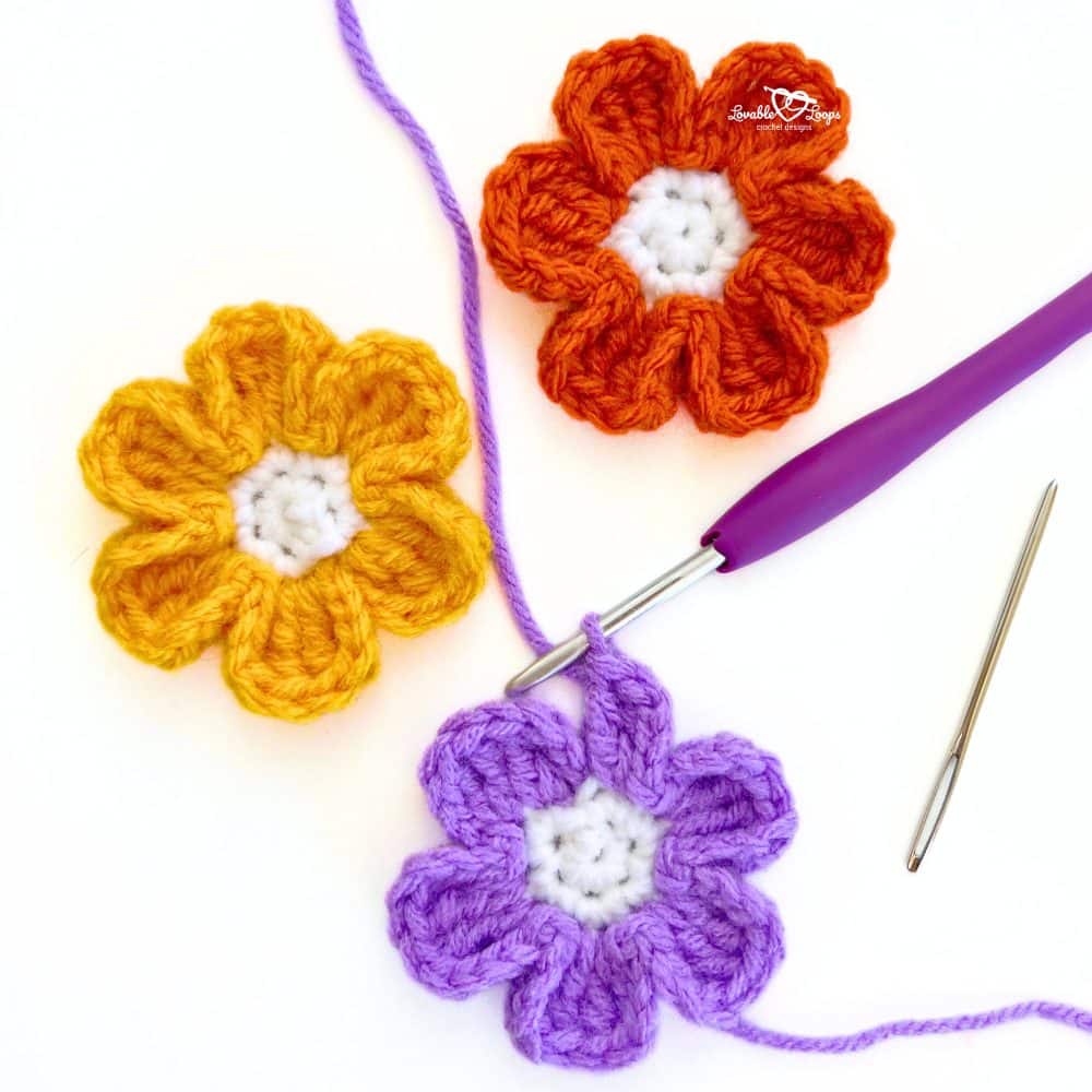 Crochet small flower pattern in progress with a purple flower on a hook, next to finished orange and yellow flowers. Easy fall crochet flower tutorial.