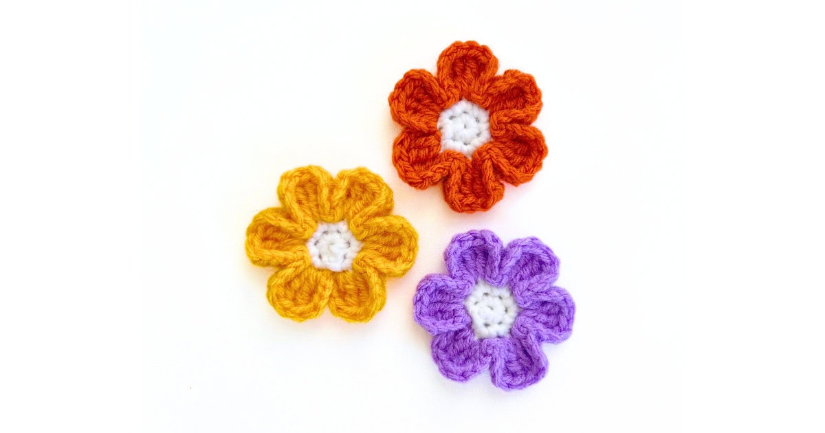 Three crochet small flowers made with a white center and colorful petals in orange, gold, and purple yarn. Easy fall crochet flower appliqué pattern, beginner-friendly.