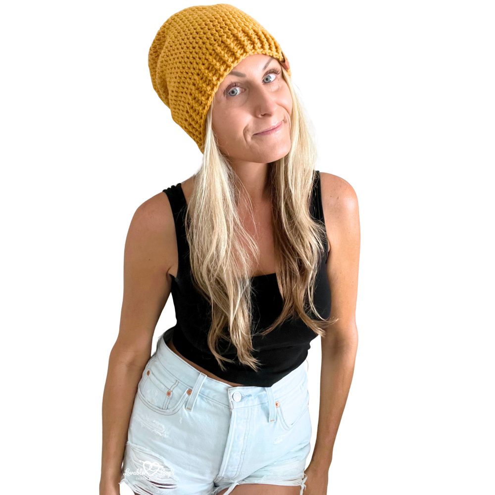 Woman wearing a mustard yellow crochet slouchy beanie and holding a teal crochet slouchy beanie, both made with a simple beginner-friendly pattern.