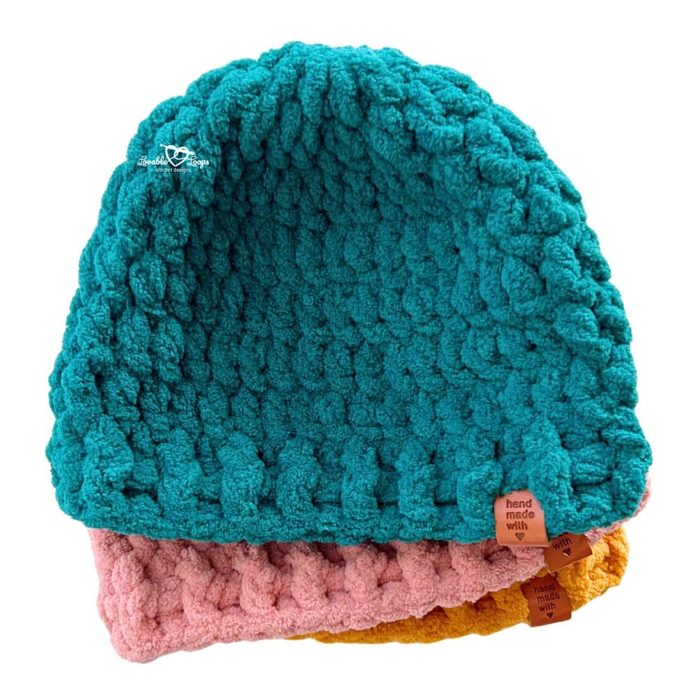 Stack of three chunky crochet beanies made with super bulky yarn, featuring teal on top, pink in the middle, and mustard yellow on the bottom, each with a ribbed brim and “handmade” tag.