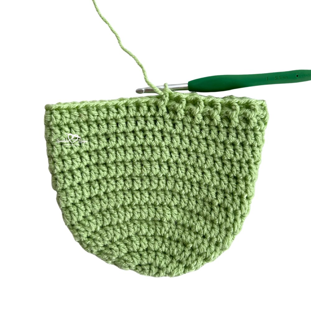 Green crochet newborn hat in progress with a 5.5 mm crochet hook still attached. Easy step-by-step crochet newborn hat pattern for beginners.