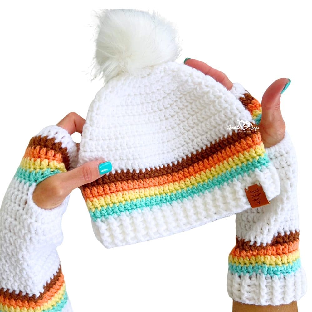 A colorful crochet pom pom beanie with bold retro stripes in brown, orange, yellow, mint, and white. The hat features a textured ribbed brim and a fluffy white faux fur pom pom on top.