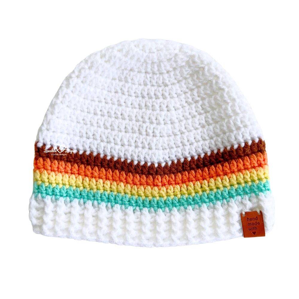 A handmade crochet beanie hat featuring bold retro-inspired stripes in white, brown, orange, yellow, and mint. The hat includes a wide ribbed brim and a leather "handmade with love" tag.