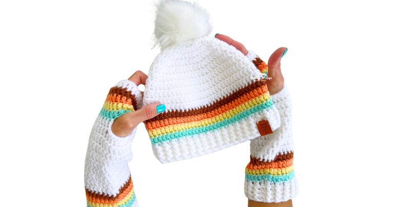 Hands wearing white crochet fingerless gloves with colorful retro stripes (brown, orange, yellow, teal) holding a matching crochet beanie with a faux fur pom pom on top. The hat and gloves both feature ribbed cuffs and bold color block accents. Great example of a coordinated handmade winter accessory set.