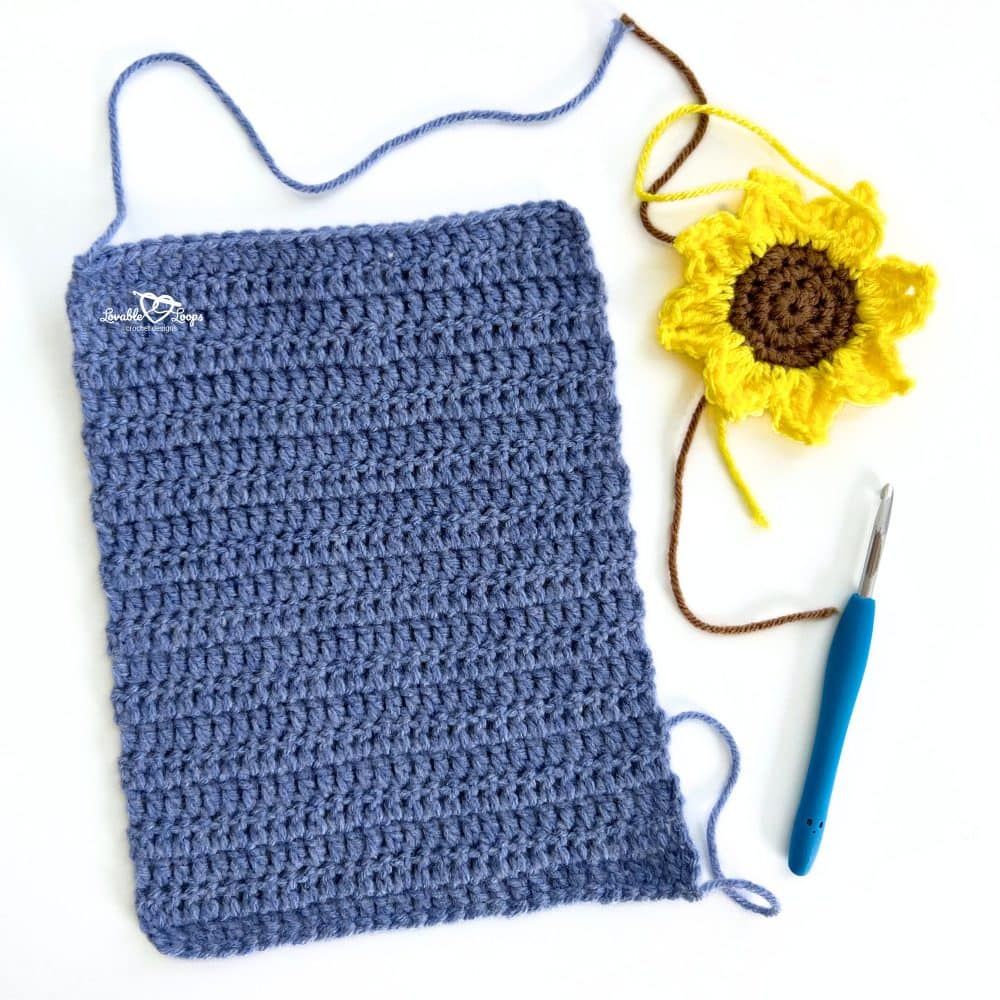 Flat blue crochet rectangle shown beside a finished yellow sunflower motif and crochet hook, step one of making crochet sunflower slippers.