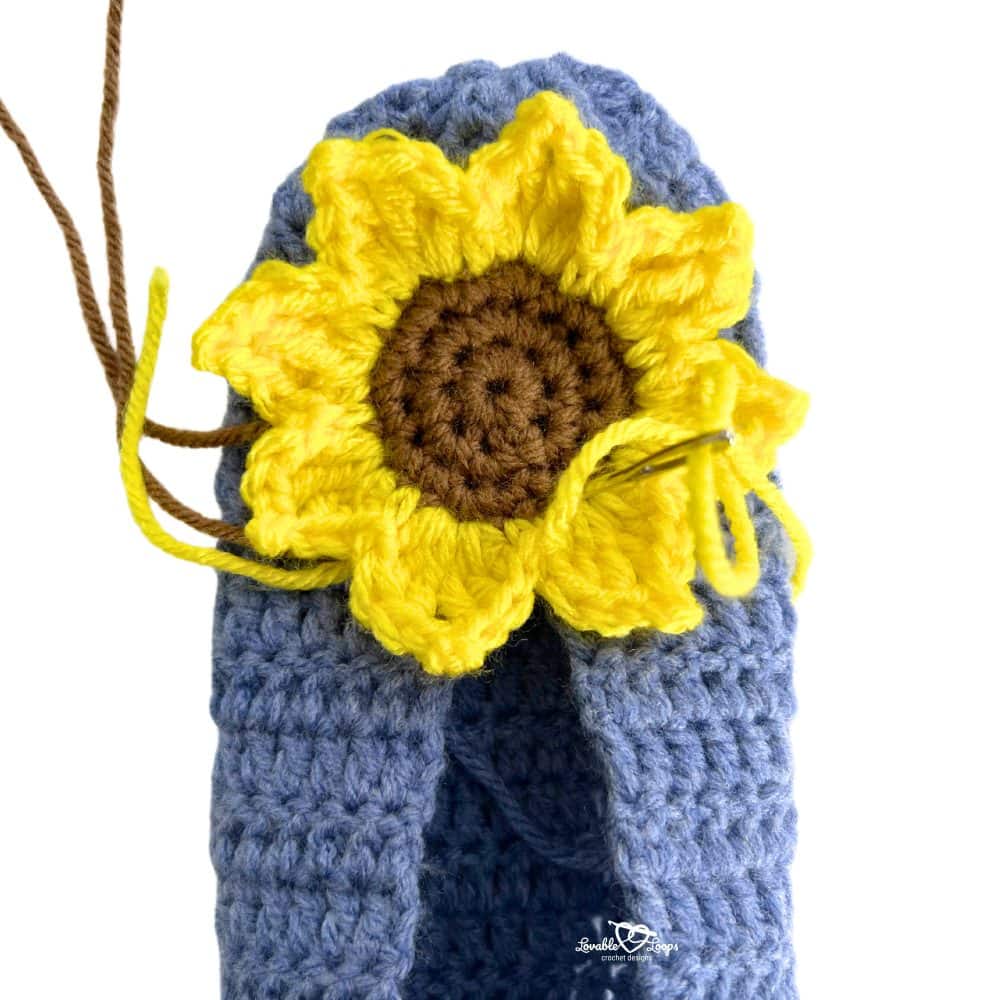 Close-up of a yellow crochet sunflower being stitched onto the top of a blue crochet slipper with a yarn needle.