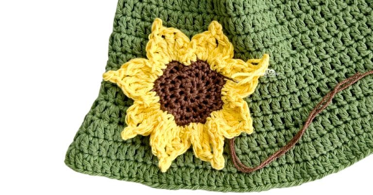 Close-up of a yellow and brown crochet sunflower applique being sewn onto a green crochet background. Free crochet sunflower pattern and easy crochet flower applique tutorial.