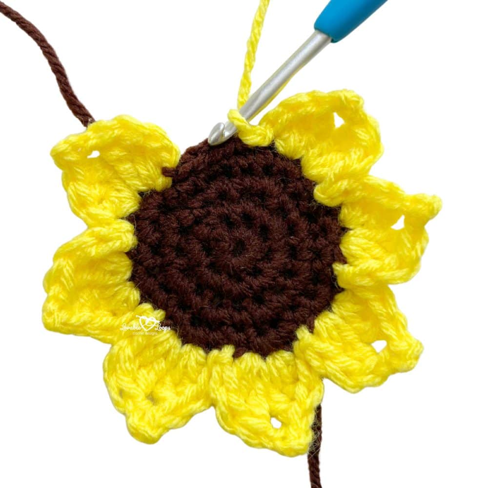 Crochet sunflower applique in progress with a blue crochet hook working into yellow petals. Step-by-step sunflower applique tutorial and free crochet flower pattern.