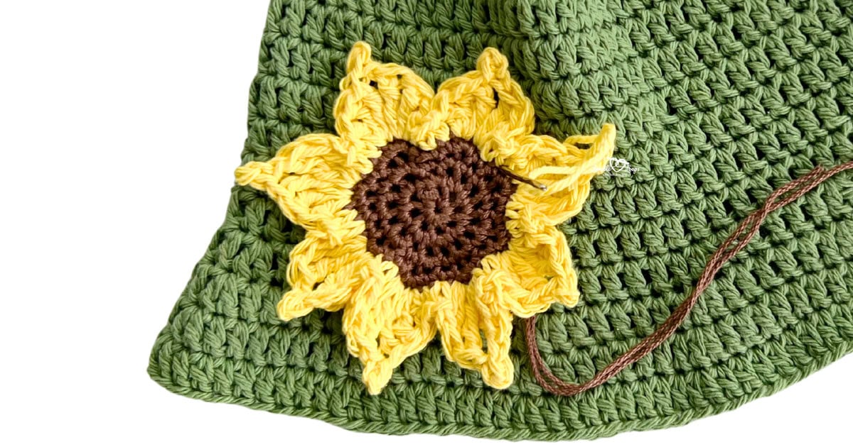 Close-up of a yellow and brown crochet sunflower applique being sewn onto a green crochet background. Free crochet sunflower pattern and easy crochet flower applique tutorial.