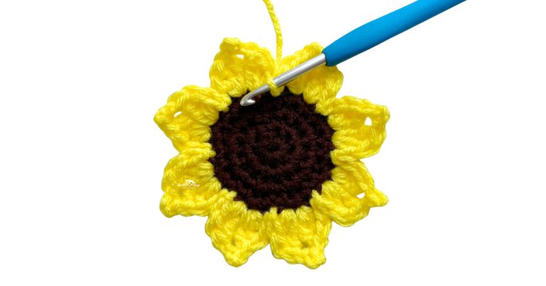 Close-up of a crochet sunflower applique in progress with yellow petals and a brown center. A blue crochet hook is shown working into the stitches. Free crochet sunflower pattern and easy flower applique tutorial for beginners.