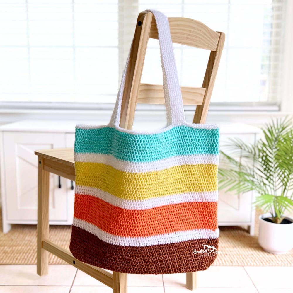 Crochet tote bag with bold, colorful stripes hanging neatly on the back of a wooden chair. The bag features wide stripes in teal, yellow, orange, brown, and white, made using a free and beginner-friendly crochet pattern.