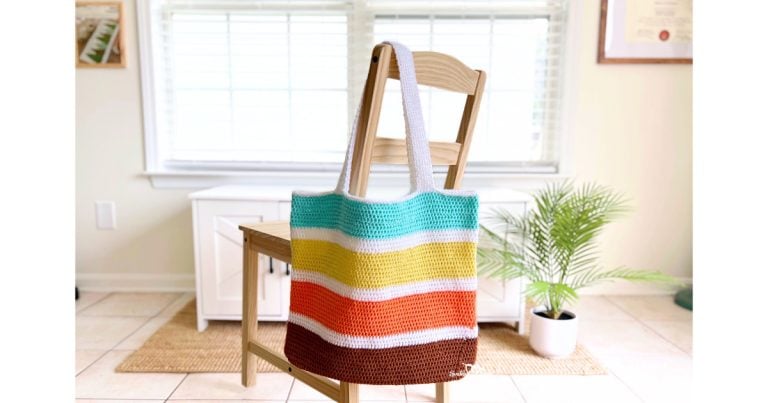 Colorful crochet tote bag with wide horizontal stripes in brown, orange, yellow, teal, and white, hanging on a wooden chair in a bright room. This handmade tote is part of a free and beginner-friendly crochet bag pattern.