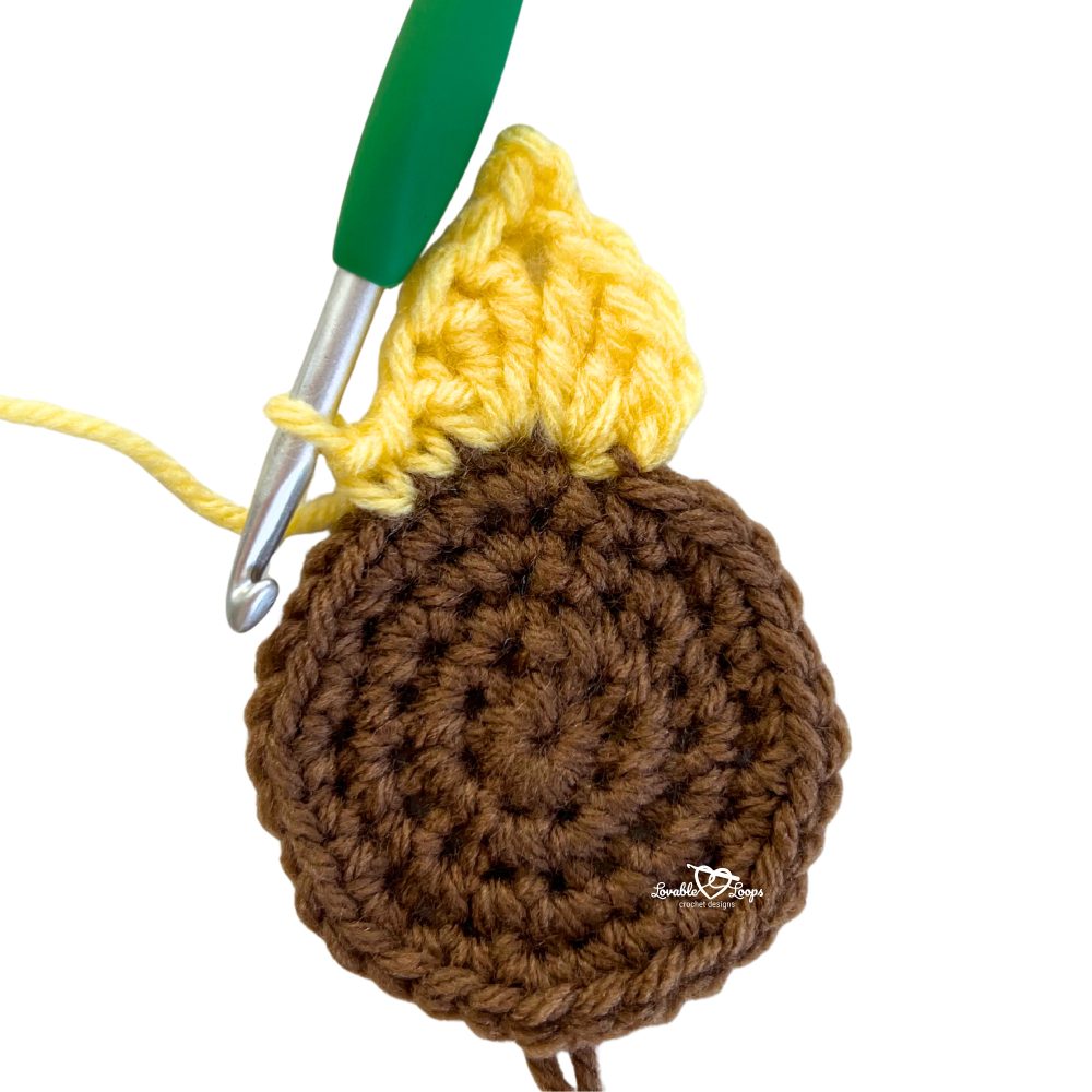 Close-up of a crochet sunflower applique in progress, showing brown center with yellow petals being worked using a crochet hook.