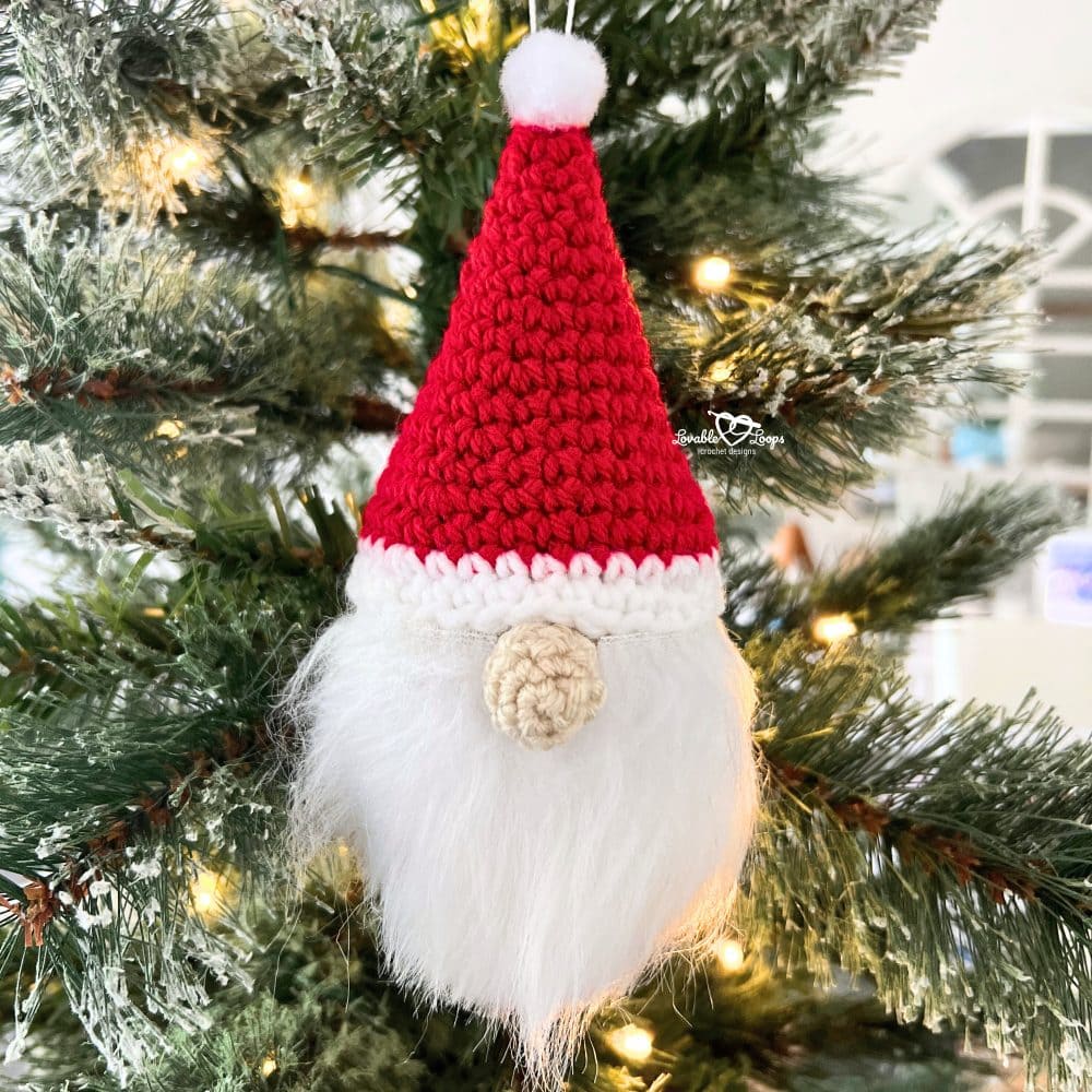 Handmade crochet Christmas gnome ornament with red hat, white trim, faux fur beard, and pom pom, hanging on a decorated Christmas tree.