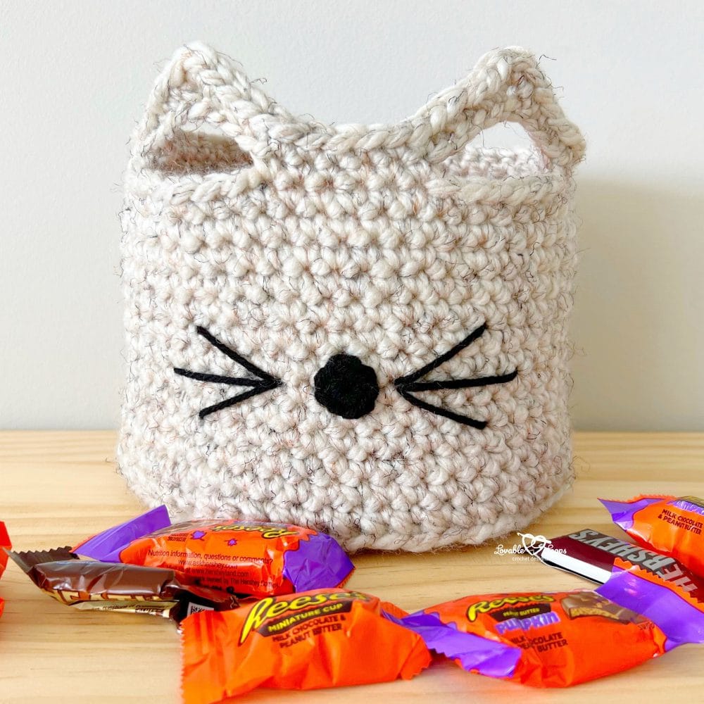 Handmade crochet cat ear basket in neutral yarn with black embroidered whiskers and nose, styled with candy around it. Perfect for Halloween candy, craft supplies, or small toy storage.