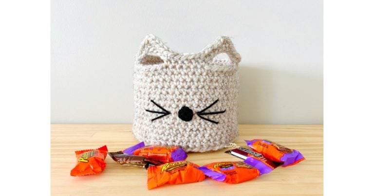Crochet basket with pointy cat ears, nose, and whiskers, sitting on a wooden table surrounded by Halloween candy. A free crochet cat basket pattern that’s fun, cute, and beginner-friendly.