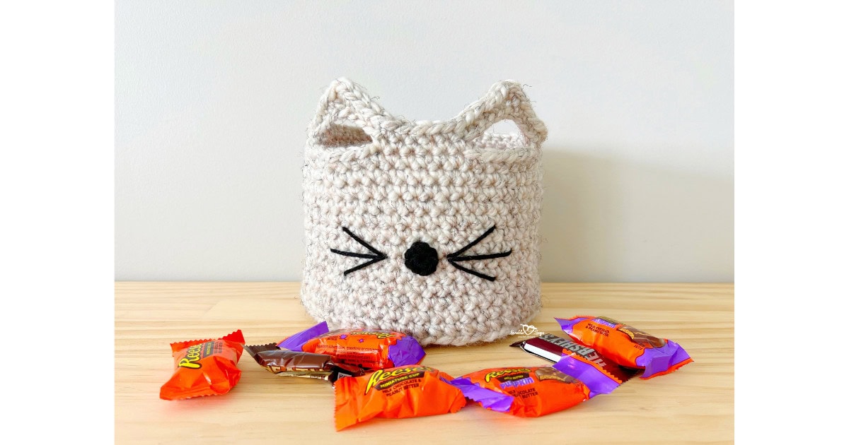 Crochet basket with pointy cat ears, nose, and whiskers, sitting on a wooden table surrounded by Halloween candy. A free crochet cat basket pattern that’s fun, cute, and beginner-friendly.