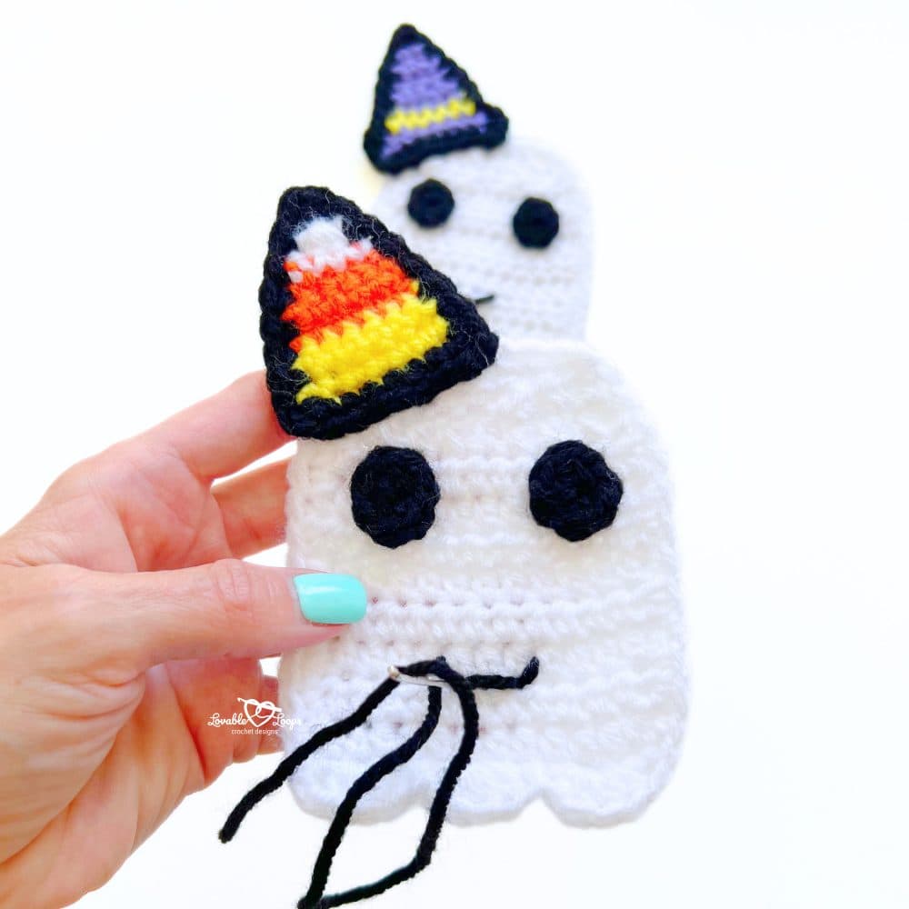 Hand holding a crochet ghost applique with a candy corn hat, while another ghost with a purple hat is shown in the background. Beginner crochet ghost pattern.