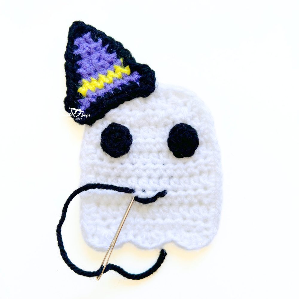 Crochet ghost applique with a purple witch hat being stitched with a yarn needle to add the smile. Step-by-step crochet ghost applique pattern.