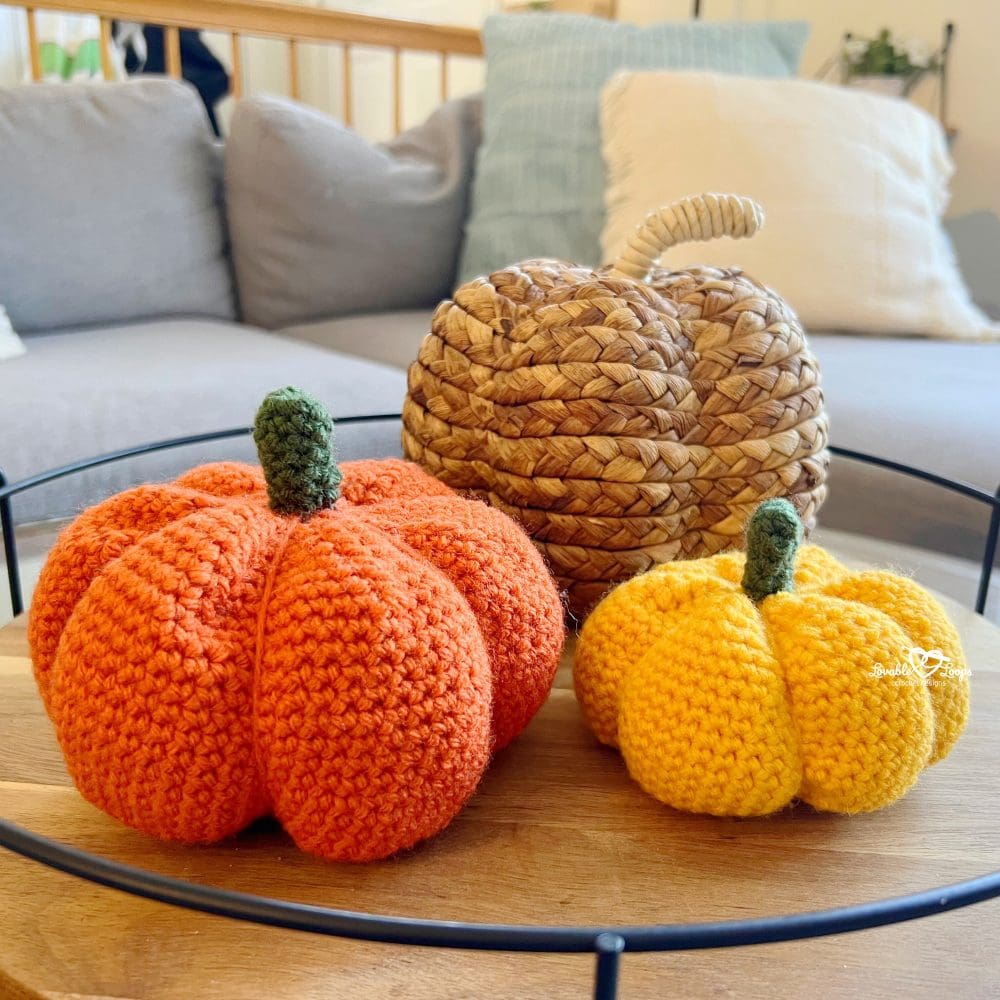 Handmade crochet pumpkins in orange and yellow yarn with green stems displayed on a wooden tray in a cozy living room. Beginner crochet pumpkin pattern in two sizes, perfect for autumn decorating.