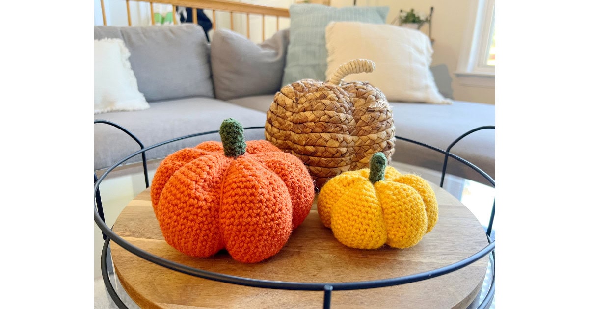 Orange and yellow crochet pumpkins with green stems displayed on a wooden tray in a cozy living room. Easy beginner crochet pumpkin pattern in two sizes, perfect for fall decorating.