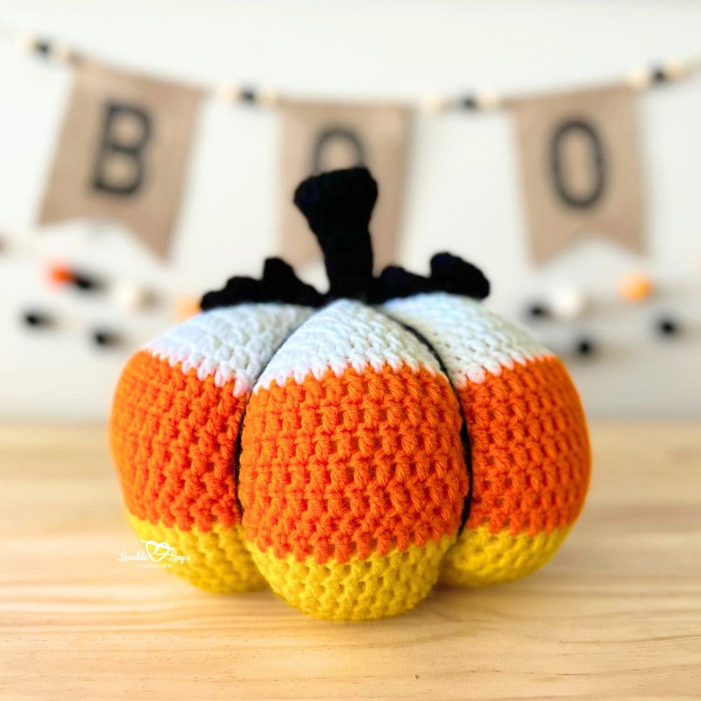 Handmade crochet pumpkin in candy corn colors of yellow, orange, and white, styled with a black stem and Halloween décor in the background.