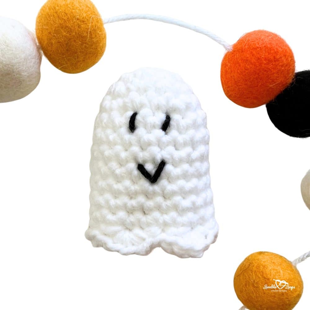 Small crochet mini ghost with stitched black eyes and mouth, hanging on a colorful Halloween pom pom garland with orange, black, and white felt balls.