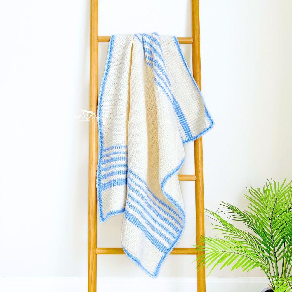 Handmade moss stitch baby blanket in cream with blue edging, draped on a wooden ladder. An easy baby boy crochet blanket pattern for beginners.
