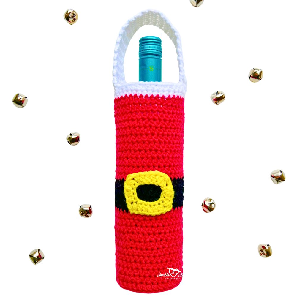 Handmade crochet Santa wine bottle cover in red with a black belt and yellow buckle, holding a teal wine bottle, surrounded by gold jingle bells. Perfect DIY Christmas wine bottle holder crochet pattern.