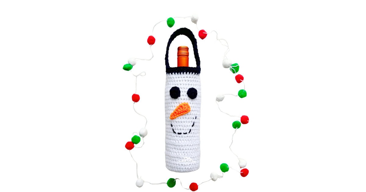 Crochet snowman wine bottle cover with black handle, carrot nose, and stitched smile, surrounded by red, green, and white holiday garland. A festive Christmas wine bottle holder crochet pattern.