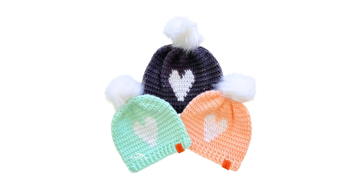 Three handmade crochet heart beanies with white faux fur pom poms in mint, peach, and gray. Each beanie features a white heart design on the front. Free crochet heart beanie pattern includes three sizes for toddler, child, and adult.