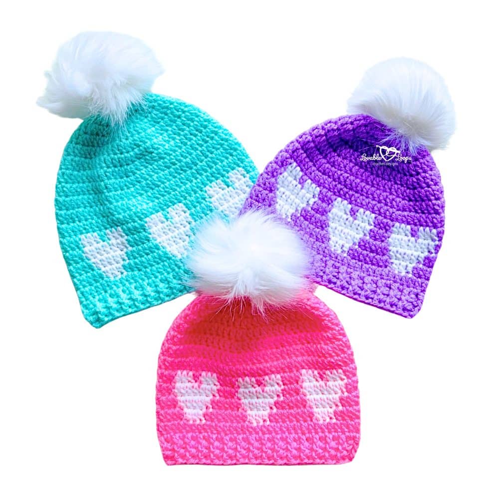 Pink, purple, and turquoise crochet heart beanies with white pom poms arranged flat. Easy crochet heart hat pattern for Valentine’s Day in three sizes with heart rows and ribbed brim.