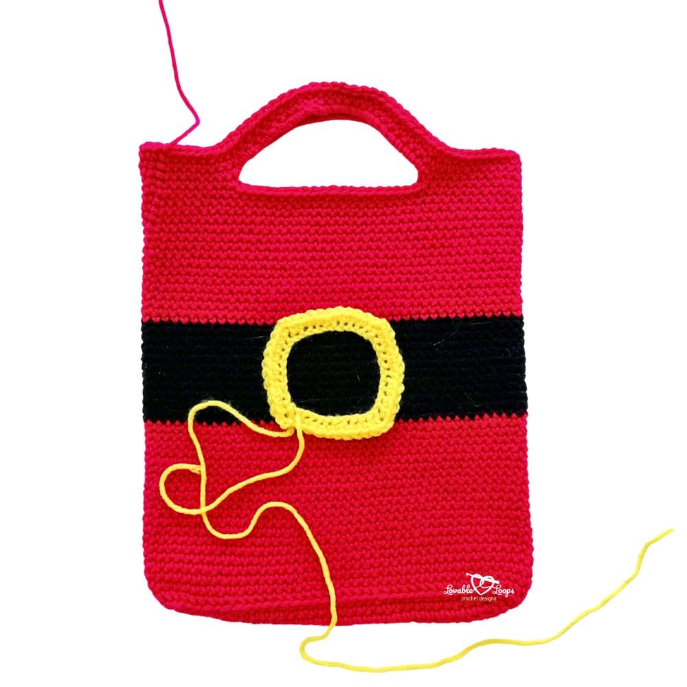 Red crochet Santa bag with a black belt stripe and yellow buckle being sewn onto the front with yarn and needle.