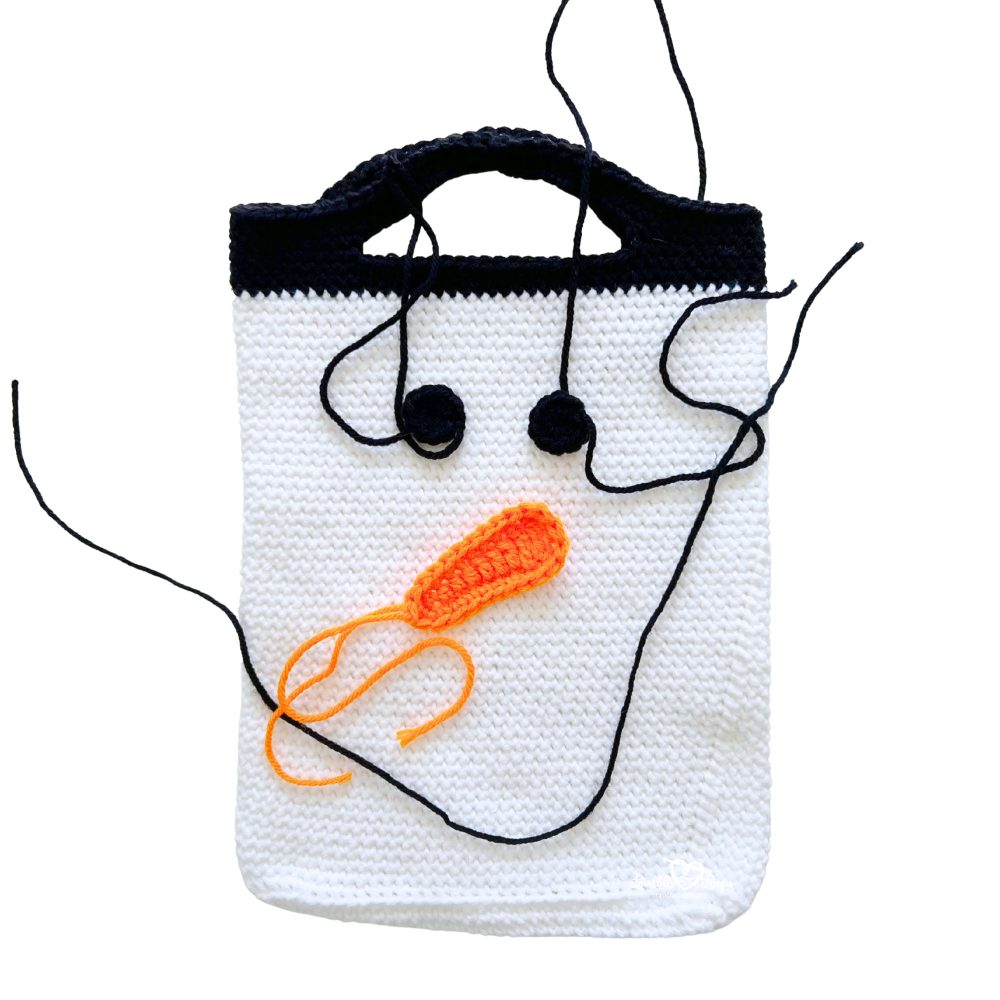 White crochet snowman bag with black handles, showing black eyes and an orange carrot nose being sewn in place.