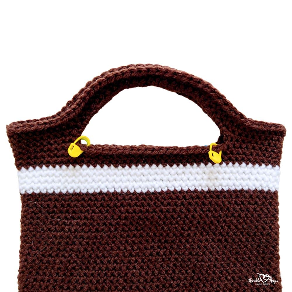 Close-up of a brown and white crochet gingerbread bag handle with stitch markers clipped near the top edge.