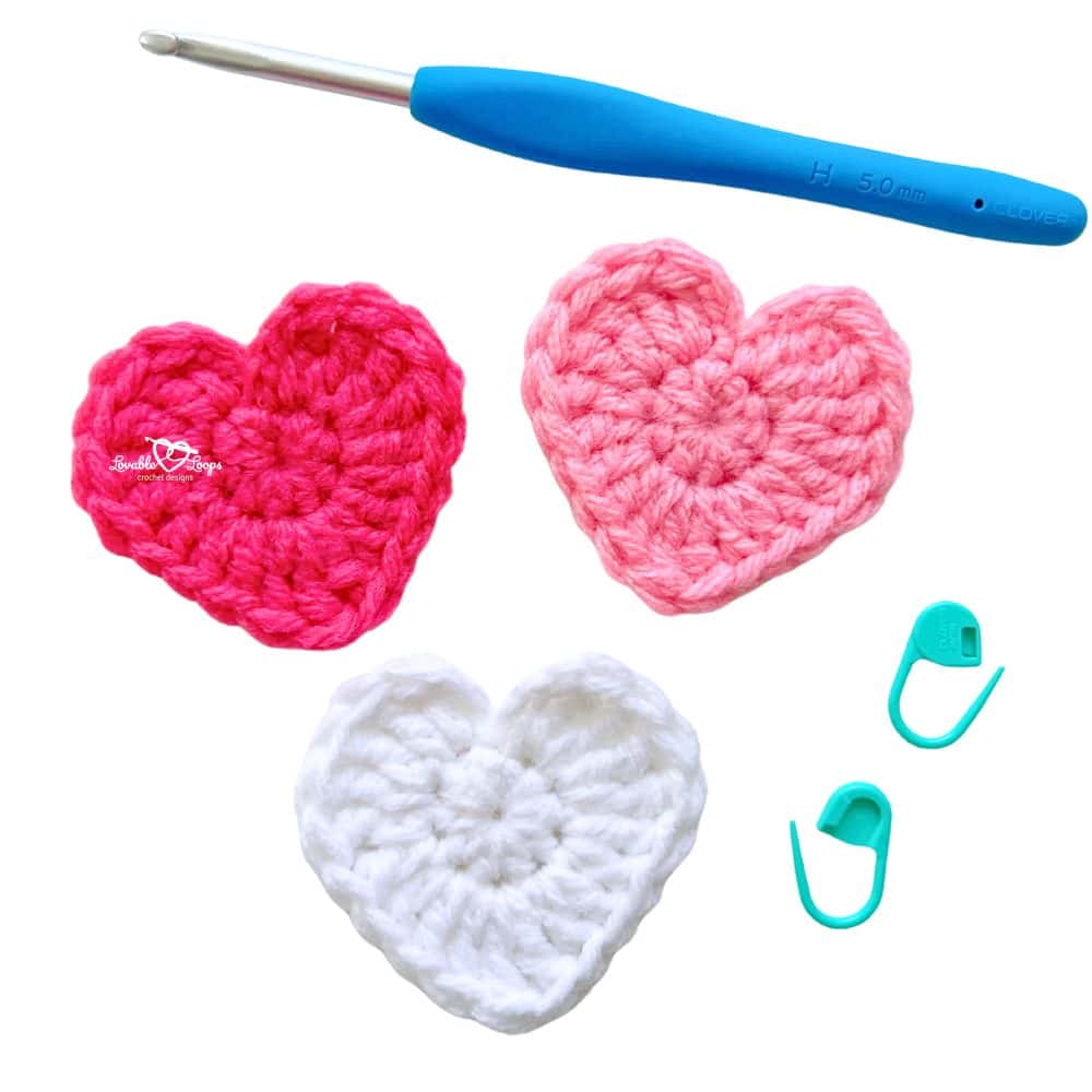 Three small crochet heart appliques in pink, red, and white shown with a 5 mm crochet hook and stitch markers. Perfect beginner project for Valentine’s Day crafts or decorations.