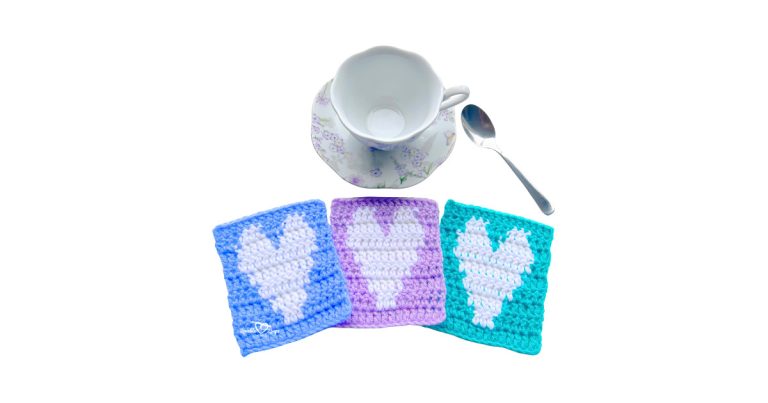 Three square crochet heart coasters in blue, purple, and teal with white hearts are arranged in front of a floral teacup and spoon, showcasing a crochet heart coaster pattern.