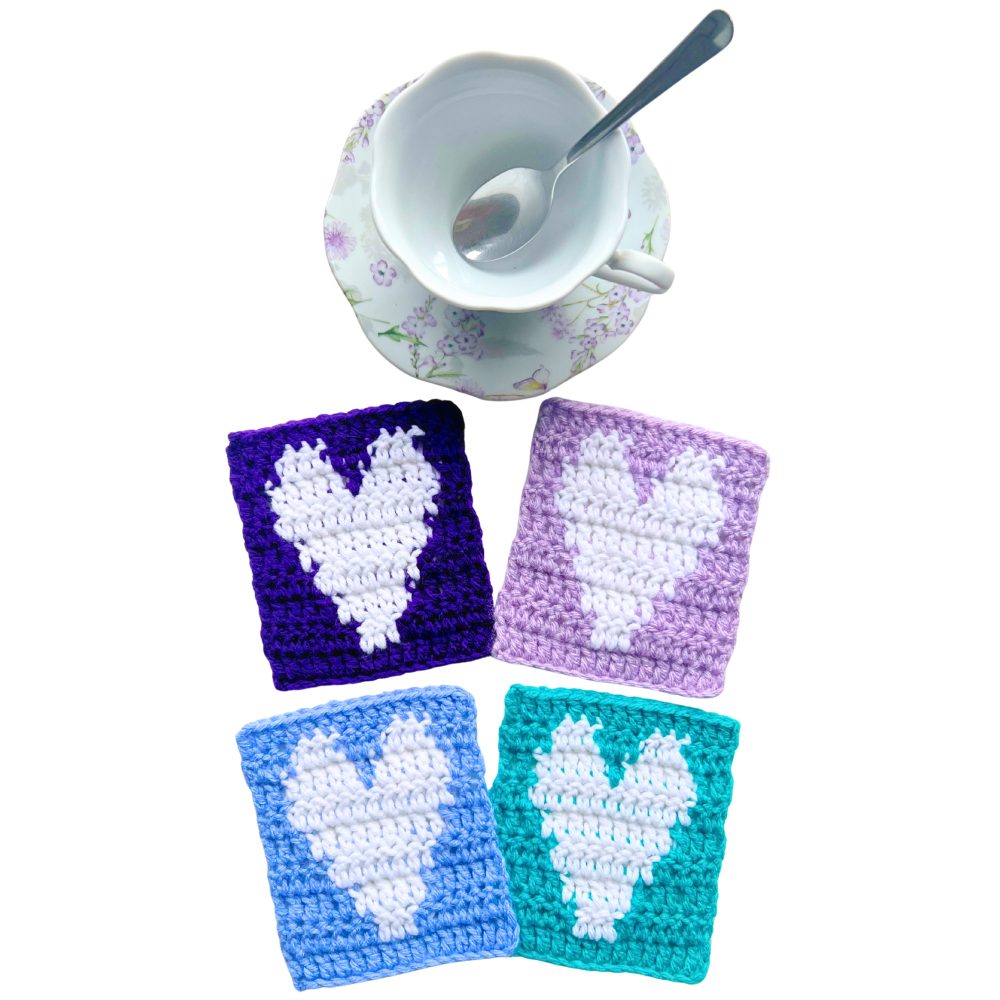 Overhead view of a floral teacup with a spoon resting inside and four crochet heart coasters in purple, lavender, blue, and teal, each with a white heart made using an easy crochet heart coaster pattern.