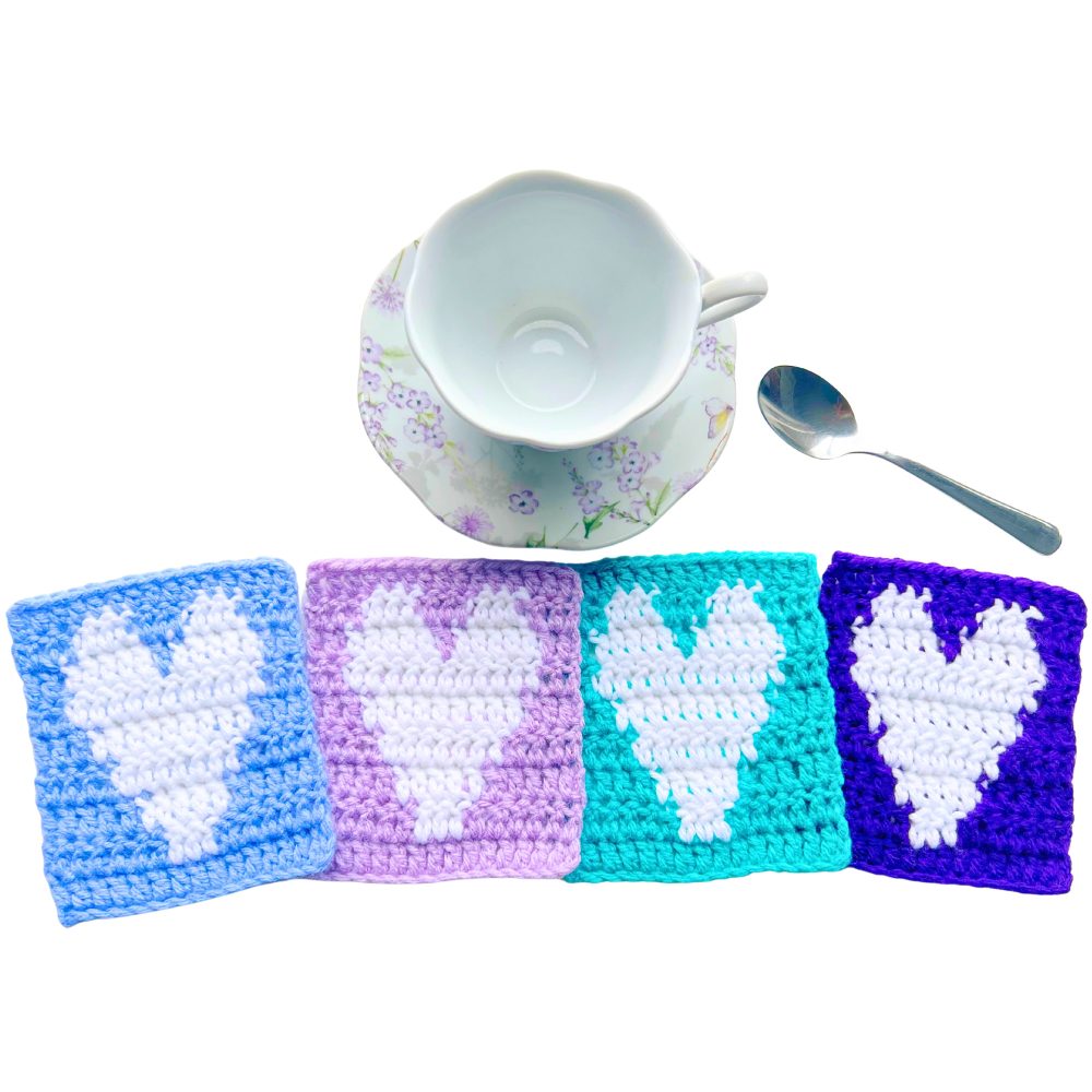 Floral teacup and saucer with a spoon beside it and four crochet heart coasters lined up in blue, lavender, teal, and purple, each worked in tapestry crochet with a white heart motif.