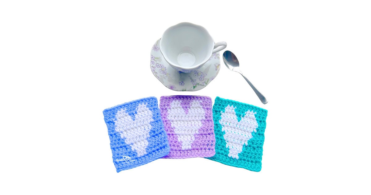 Three square crochet heart coasters in blue, purple, and teal with white hearts are arranged in front of a floral teacup and spoon, showcasing a crochet heart coaster pattern.