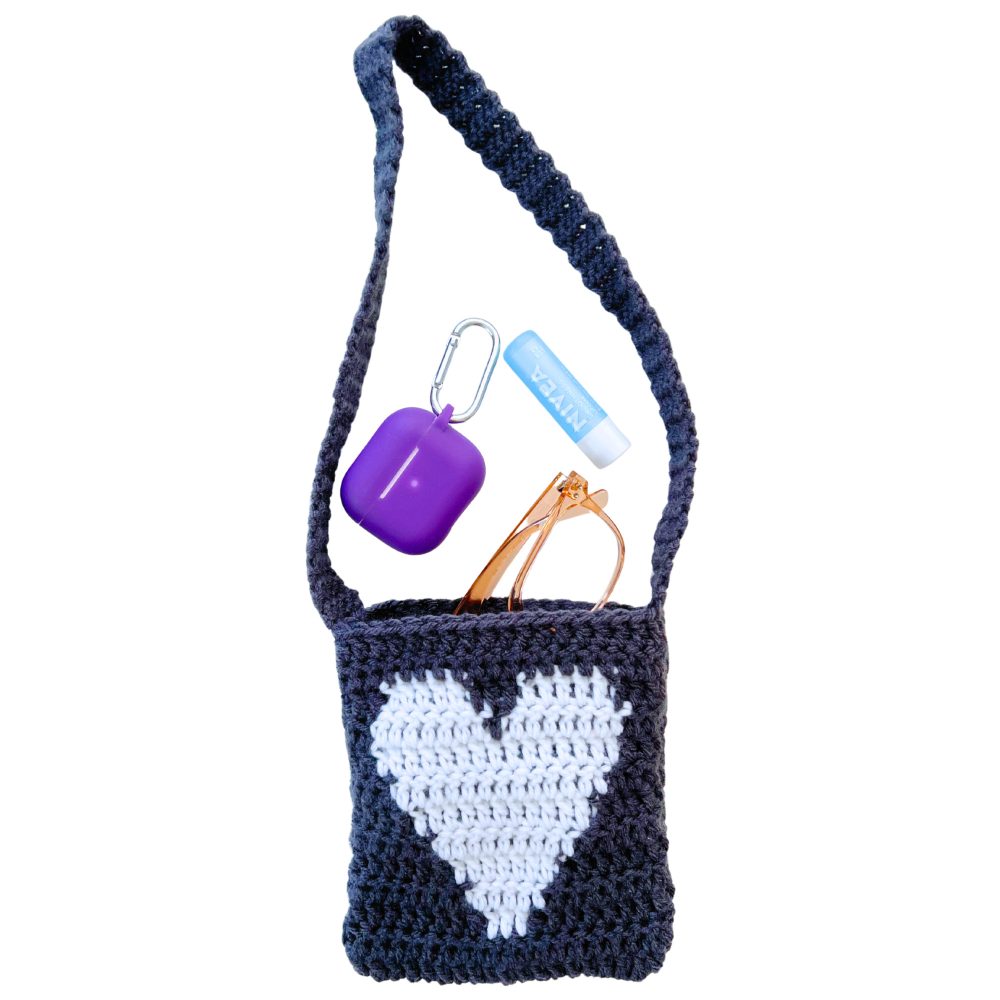 Dark gray crochet heart purse with a white tapestry heart, shown with sunglasses, a purple AirPods case, and Nivea lip balm to illustrate how much the small crochet bag can hold.