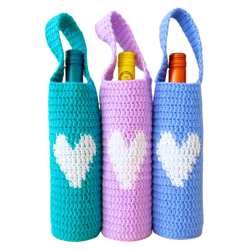 Close-up of three handmade crochet wine bottle holders with straps and large white heart designs—beginner-friendly crochet wine bottle holder free pattern and cute crochet wine bag for gifts.